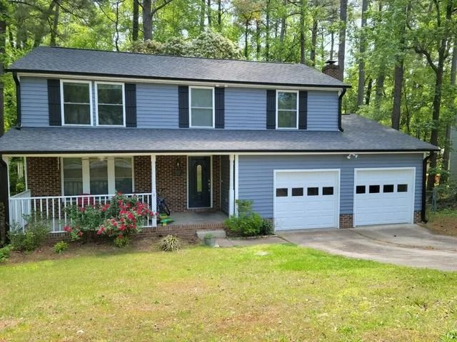 Two-story house with blue siding, white garage doors, and a porch. Red brick accents, surrounded by green trees and grass.