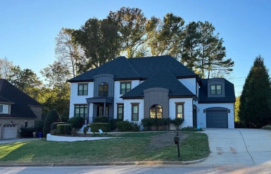 Two-story white brick house with dark roof, surrounded by greenery and trees, on a sunny day.