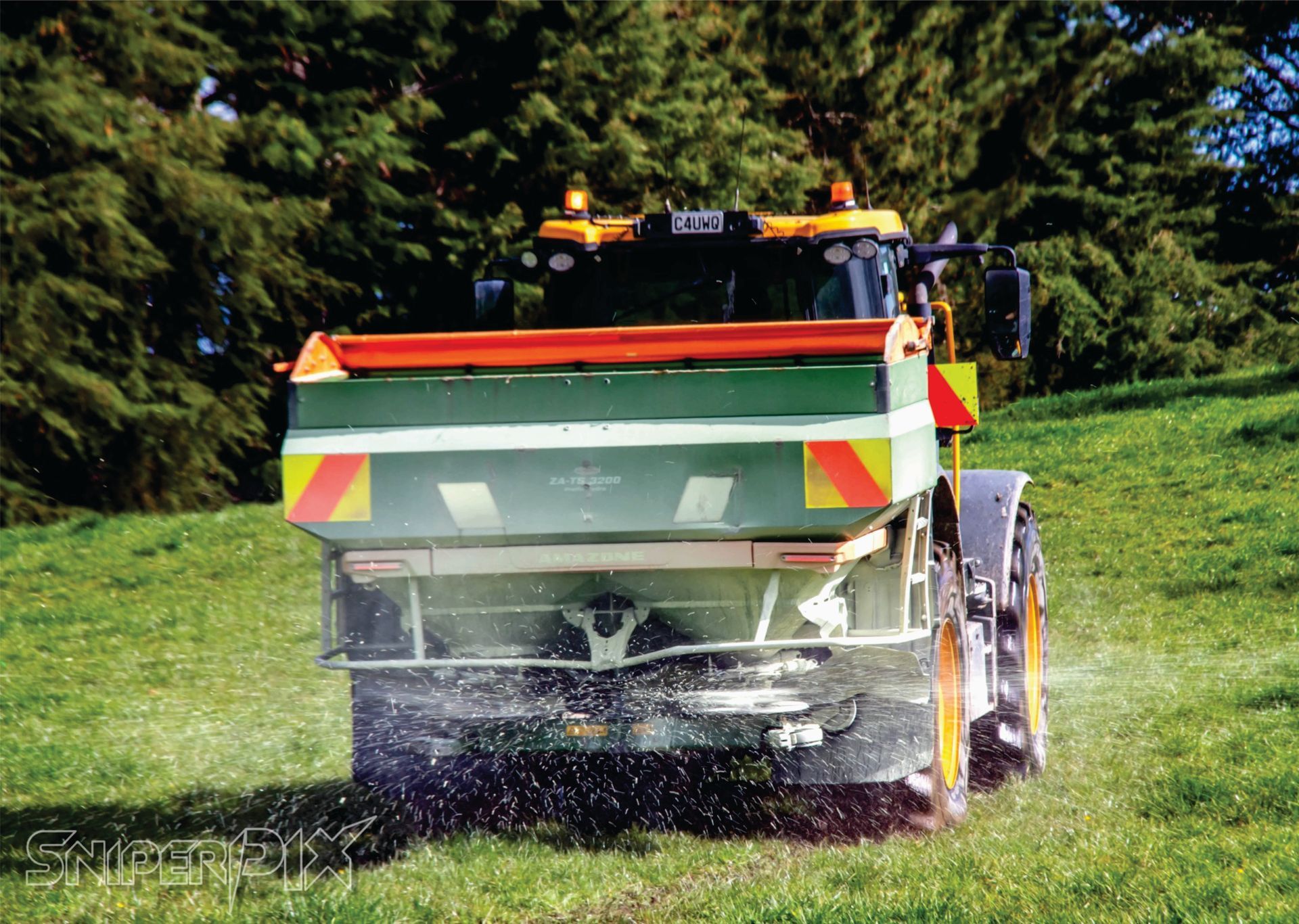 Tractor spreading fertilizer on a grassy hillside. Green and orange equipment with reflective markers.