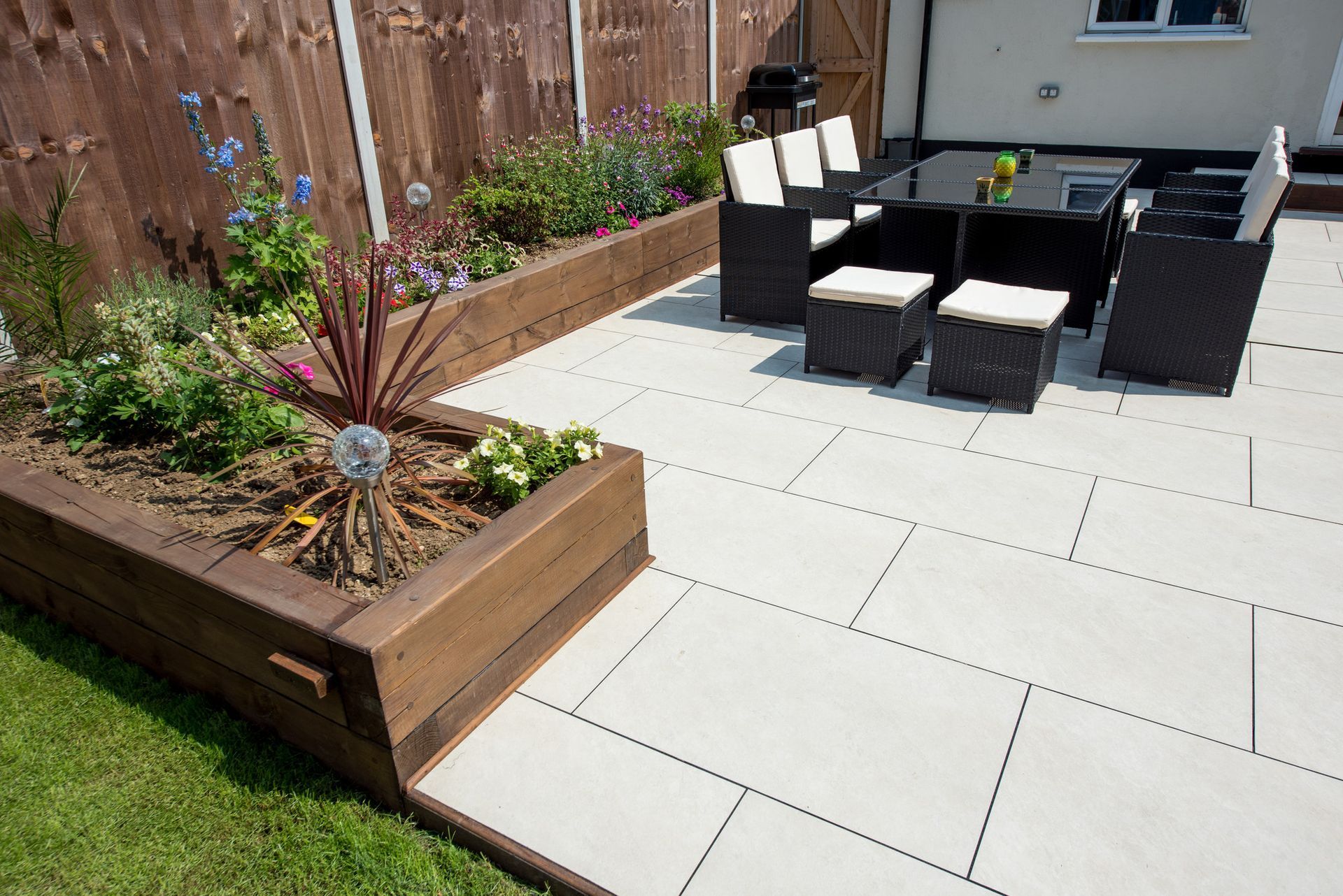 A general view of a back garden patio with white porcelain slabs, tiles, paving and wooden railway sleeper flowers beds, black rattan table and chairs with cream cushions on a bright sunny day in summer