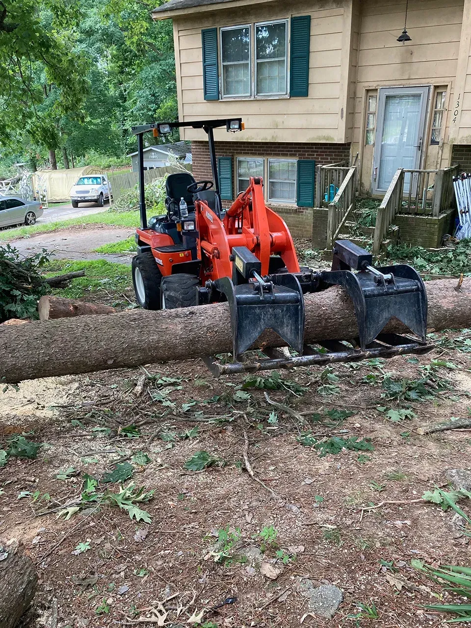 A tractor is carrying a large log in front of a house.