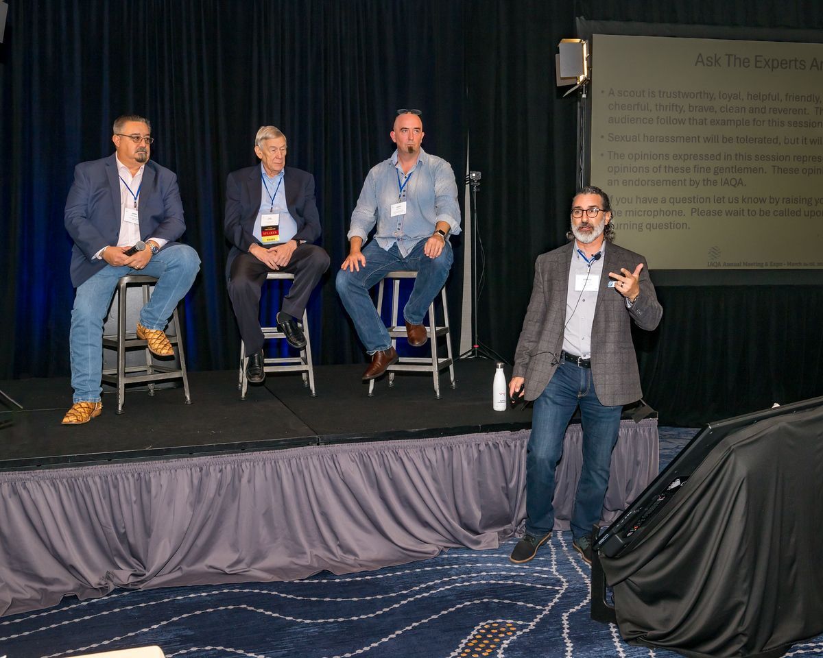 A group of men are sitting on stools on a stage while a man stands on stage giving a presentation.