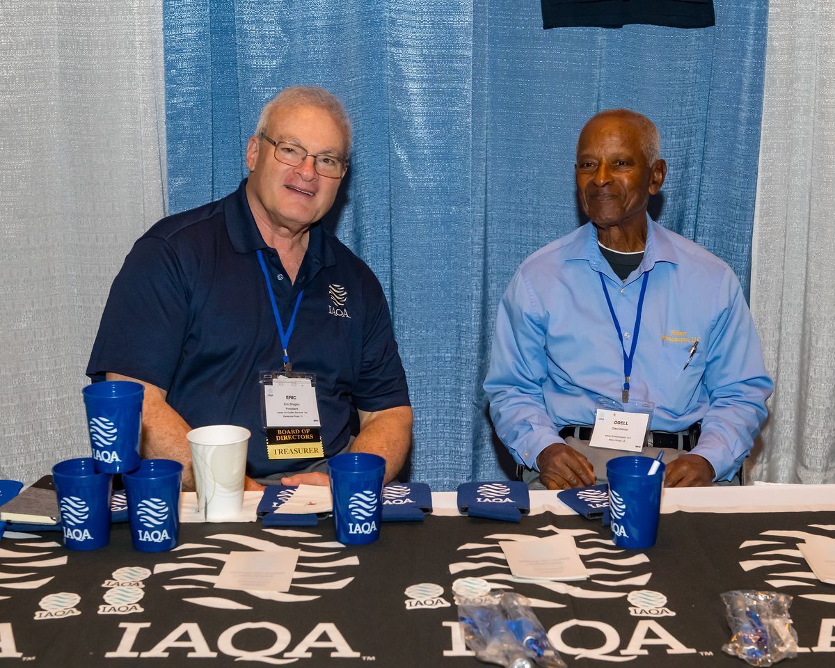 Two men sit at a table with a sign that says iaqa