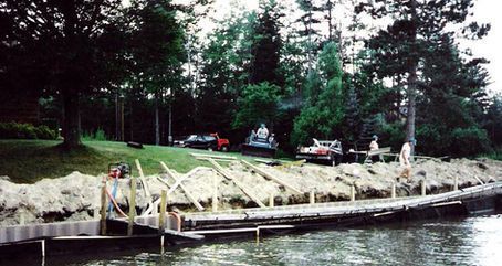 Dock construction site by a lake; workers, trucks, and trees are visible.