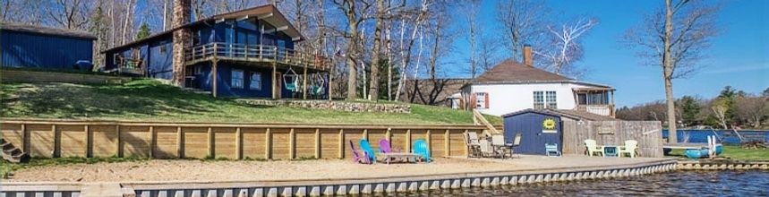 Lakeside view with blue houses, retaining wall, beach chairs, and bare trees under a blue sky.