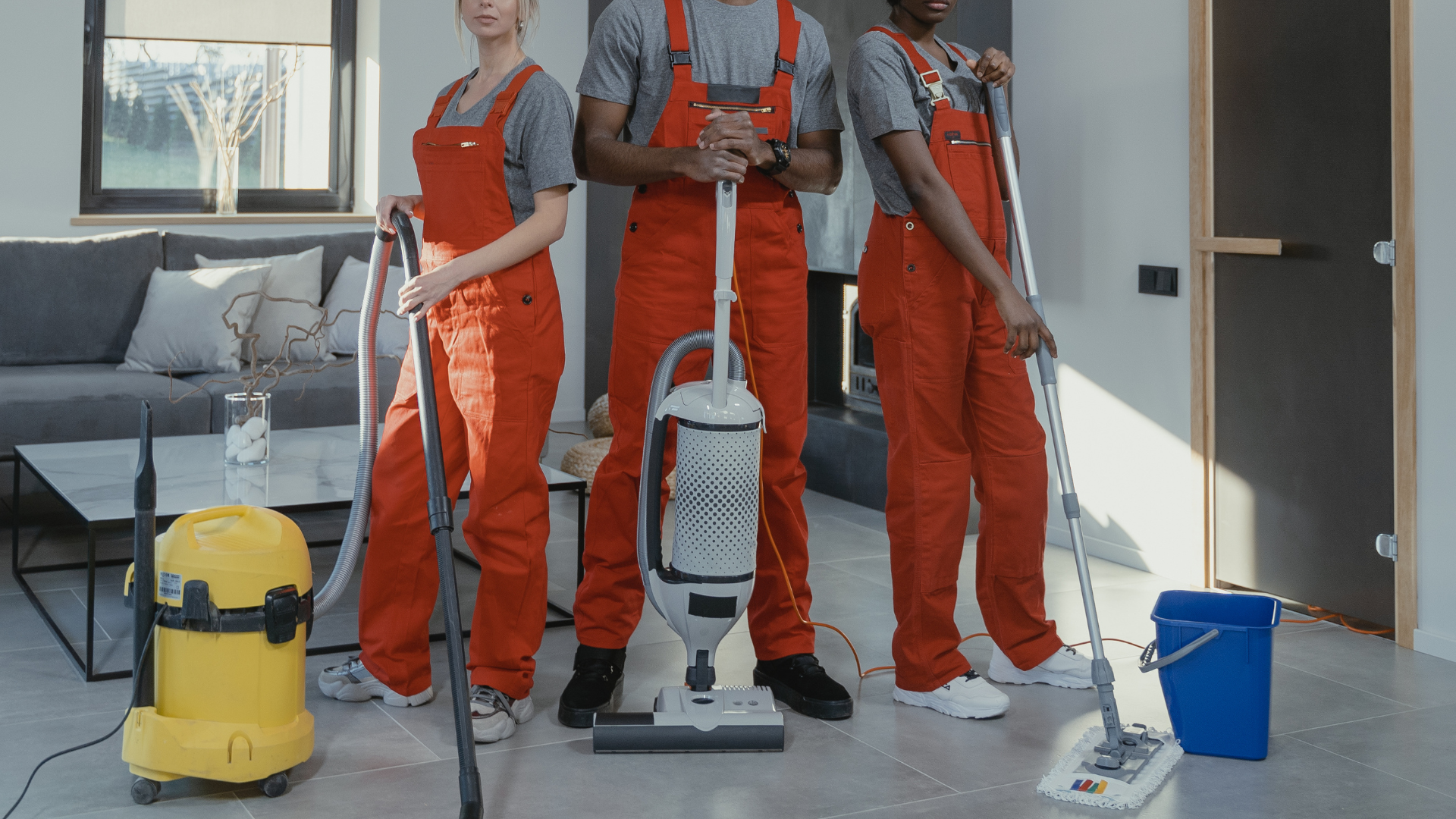 A group of cleaners are standing in a living room holding vacuum cleaners and mop.