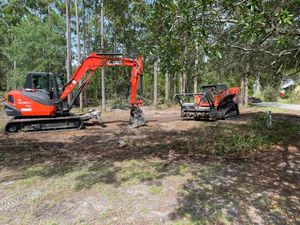 A red and black excavator is sitting in a dirt field next to a stump grinder.
