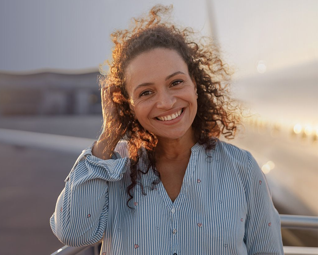 Woman with curly hair smiling outdoors, near an airplane, wearing a striped shirt.