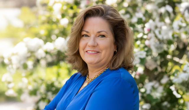 Woman sitting on a beige couch, smiling, resting hand on chin. Wearing a light blue shirt. Indoor setting.