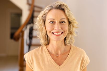 Woman with blonde hair smiles at the camera, wearing a light-colored top, staircase in background.