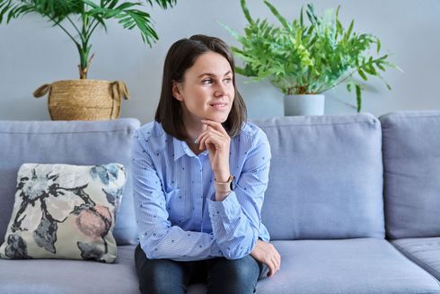 Woman sits on a sofa, hand on chin, looking away thoughtfully. Plants in background.