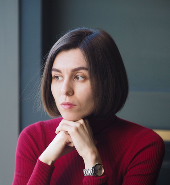 Woman with short brown hair, resting chin on hands, wearing a red turtleneck, looking pensively to the right.