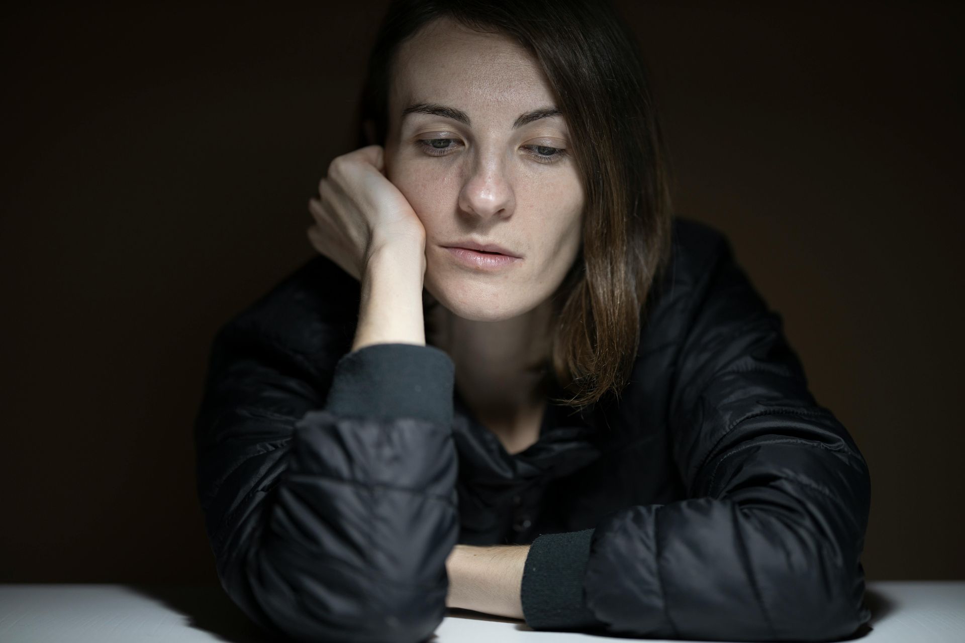 Woman with head resting on her hand, looking down with a sad expression. Dark setting.