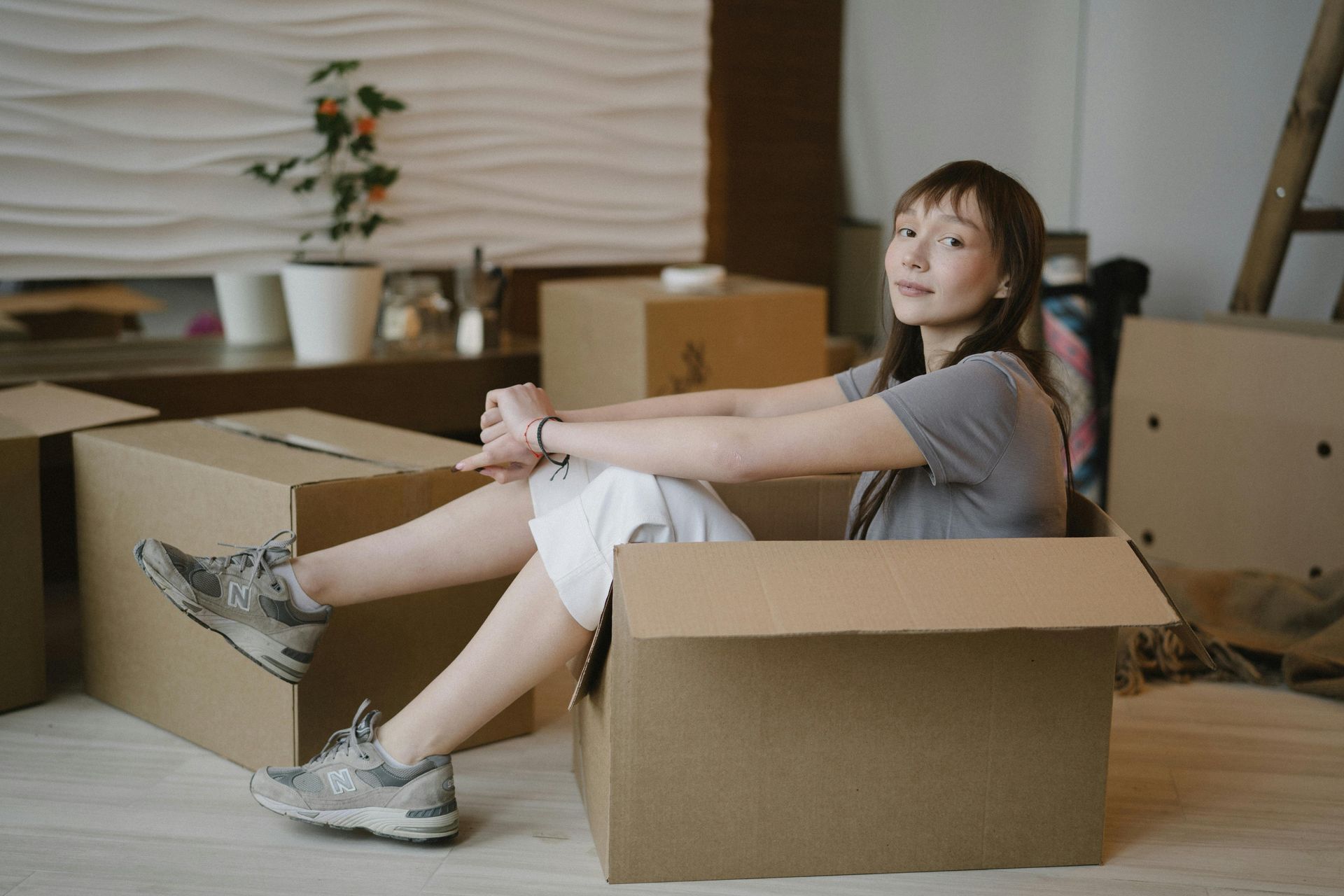 Woman sits inside a cardboard box, smiling. Boxes and a plant are in the background of the room.