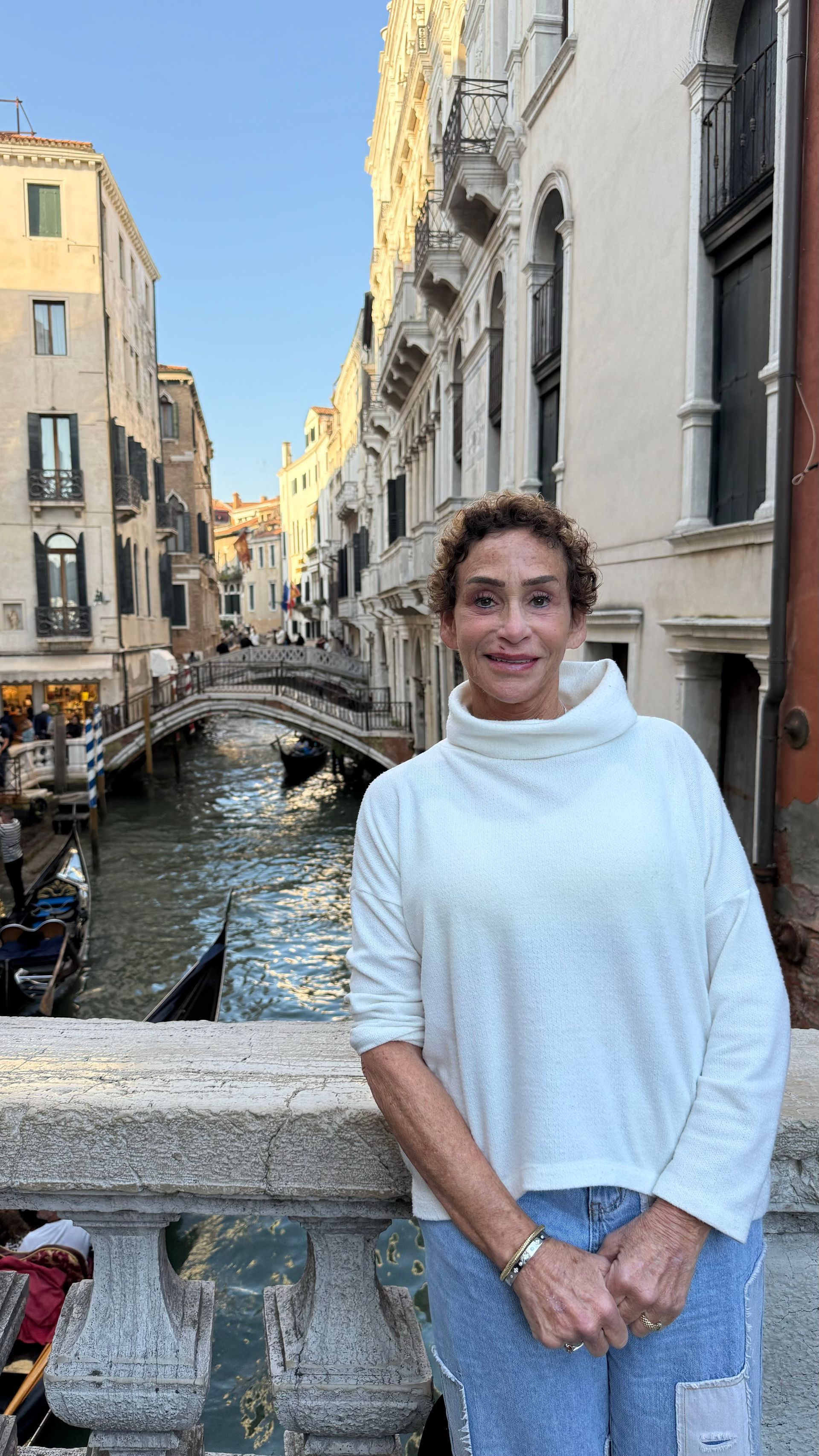 A person wearing a white sweater stands on a stone bridge overlooking a Venetian canal with gondolas and historic buildings.