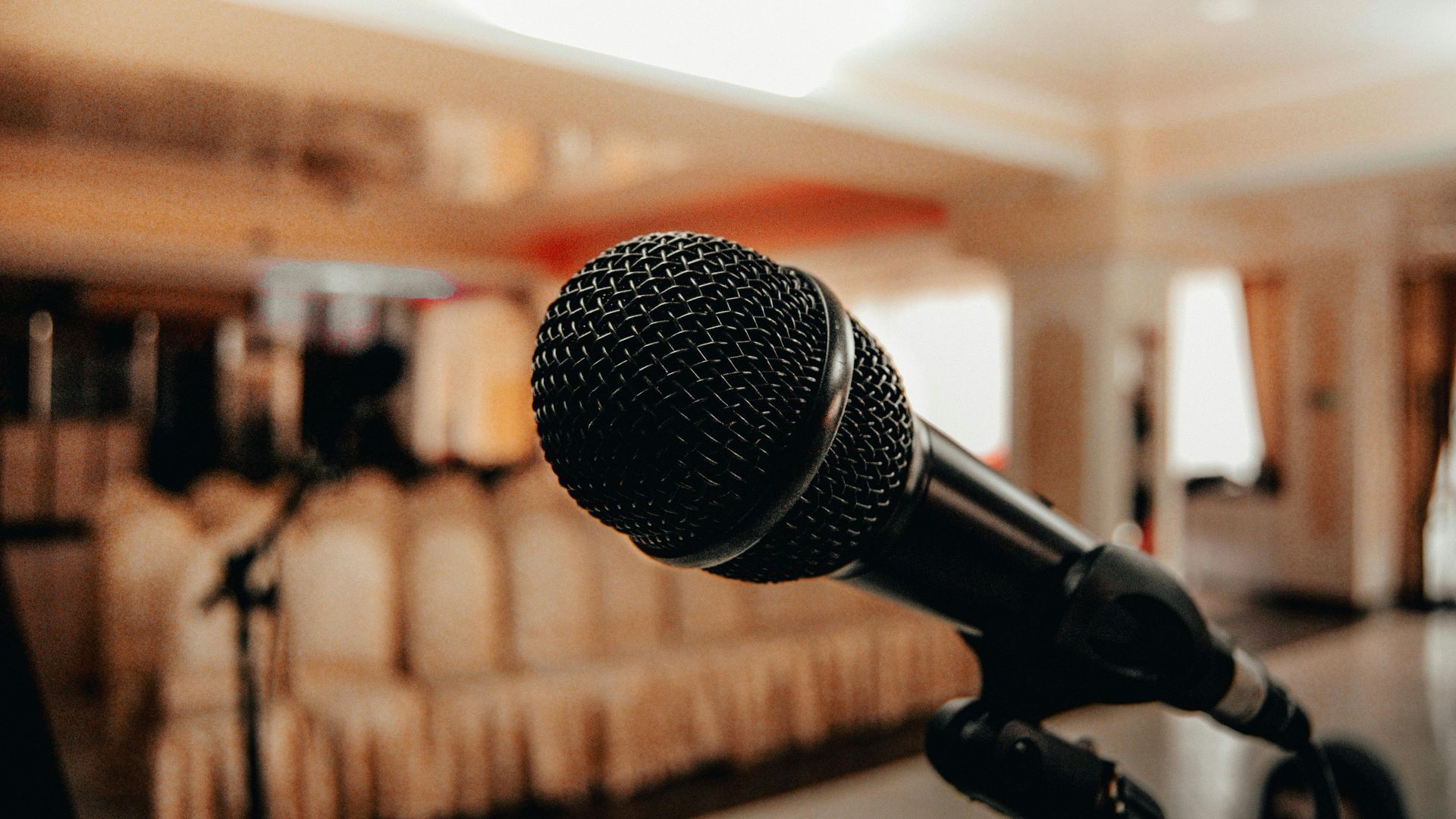 A close-up view of a black microphone on a stand, positioned in front of an out-of-focus event space with seated chairs.