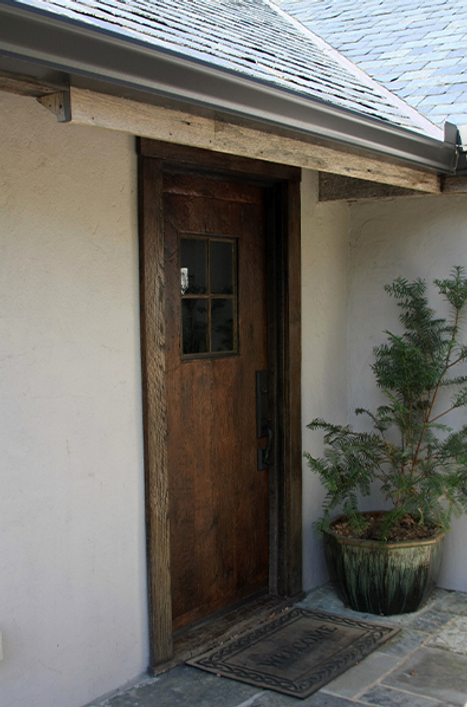 Wooden door with small window and doormat, next to potted plant, under a roof