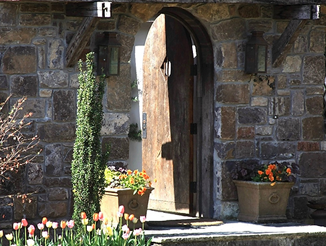 Stone building entrance with arched wooden door ajar, flanked by potted flowers, greenery, and sconces