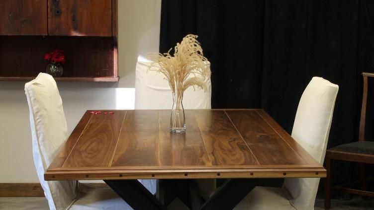 A wooden dining table with four white chairs and a vase of dried flowers, set against a dark backdrop