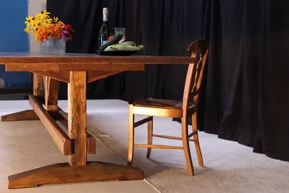 Wooden table set for one, with flowers, a bottle, and a plate of food; next to it, a chair sits against a black curtain