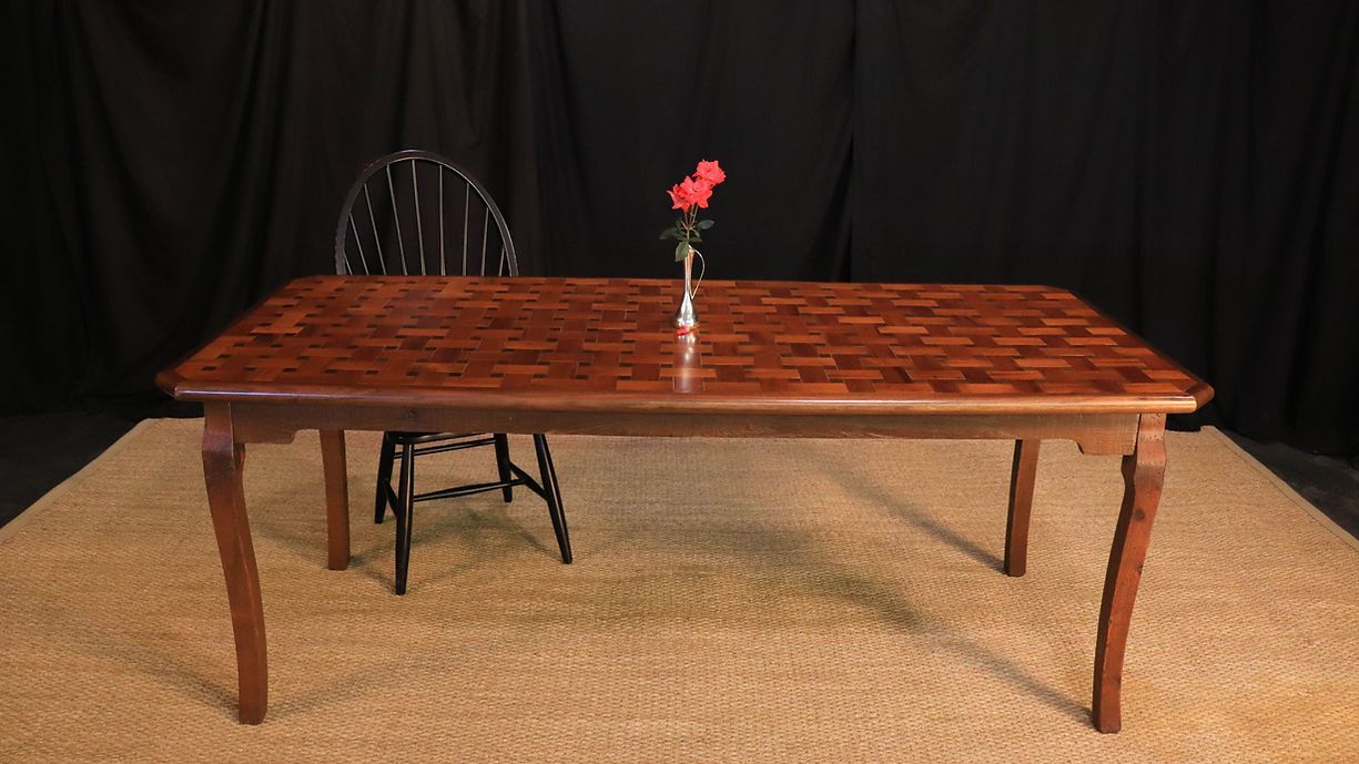 A wooden table with a mosaic top, a single chair, and a vase of red flowers against a black backdrop