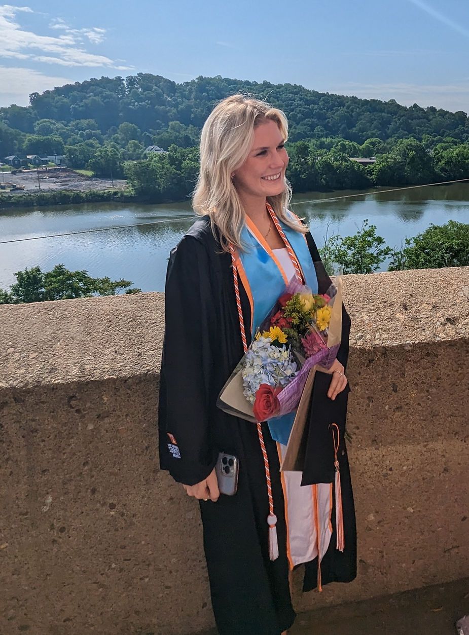 Woman in graduation gown smiles, holding flowers, overlooking a river