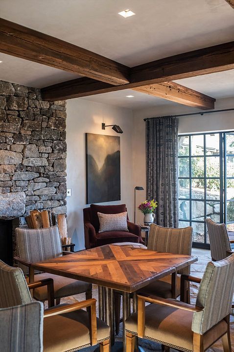 Dining area with square wooden table, patterned chairs, stone wall, wooden beams, and velvet armchair