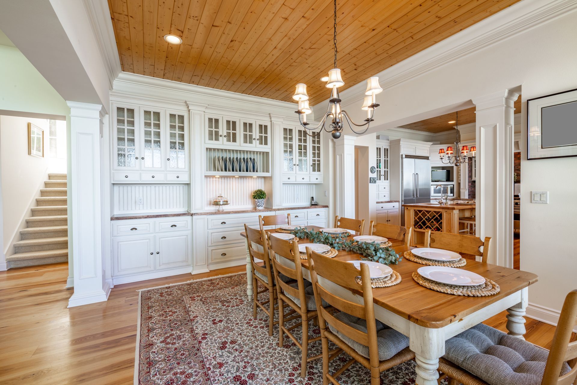 Dining room with wooden table, chairs, white hutch, wood ceiling, chandelier, rug, and stairs