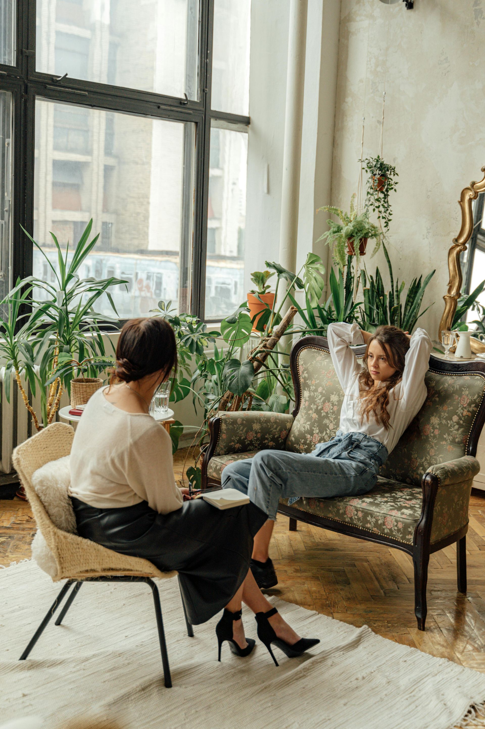 Woman in jeans on a couch with hands behind head, talking to a woman in a chair, both in a sunlit room with plants.