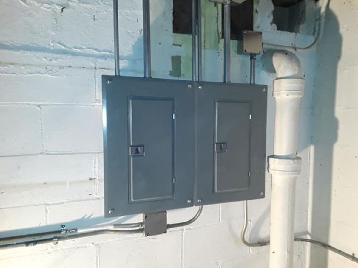 A gray electrical box is mounted on a white brick wall next to a white pipe.