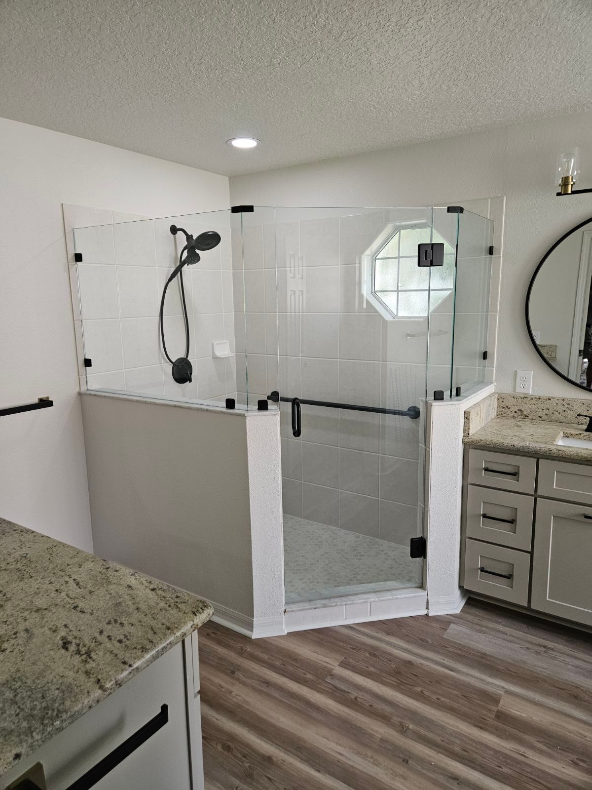 A wheelchair-accessible shower with glass doors, a handrail, and black fixtures in a bathroom with light-colored cabinets.