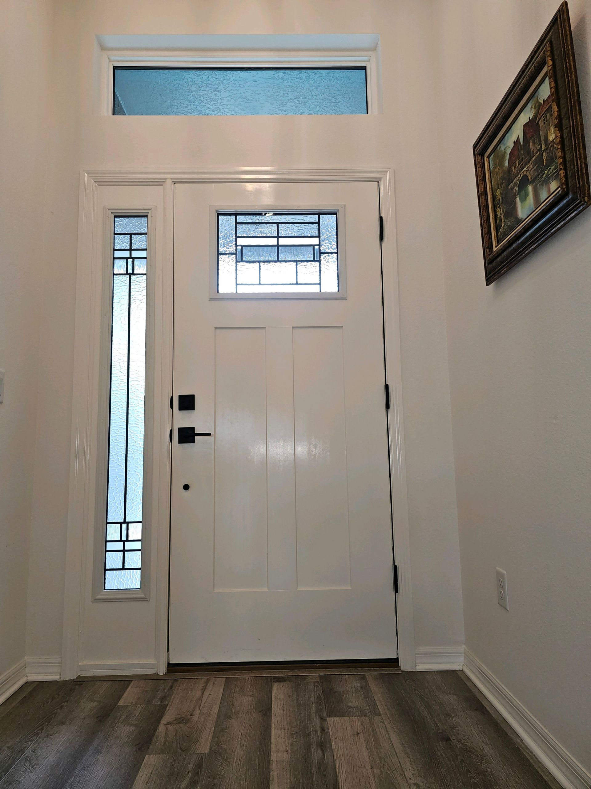 White entry door with sidelight and transom window, dark hardware, and artwork on wall.