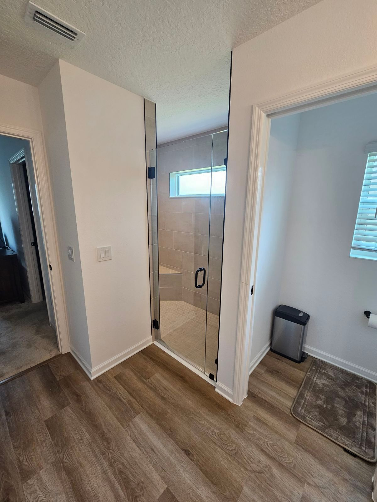 Bathroom with glass shower, beige tile, and light wood-look flooring. A trash can and toilet are also visible.