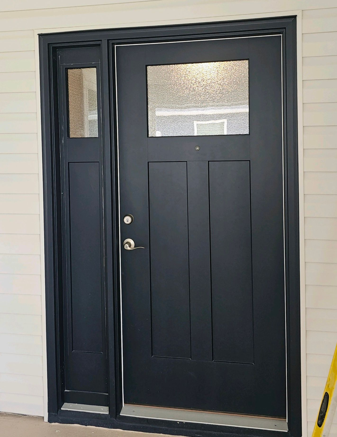 Black door with sidelight, paneled design, silver hardware, and clear textured glass. White siding background.
