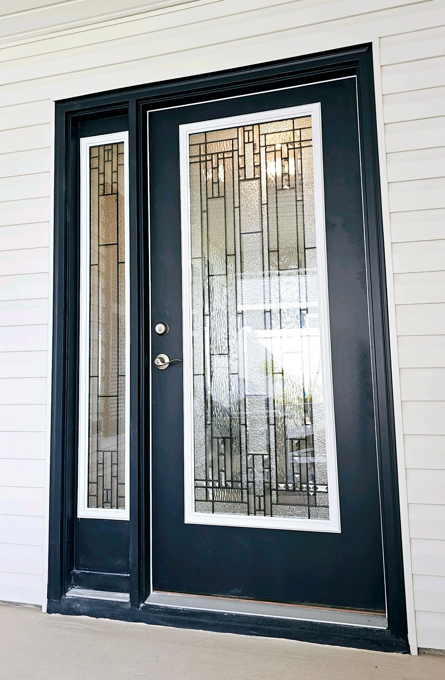 Black front door with sidelight, both with textured glass and white trim, set in a white-sided building.