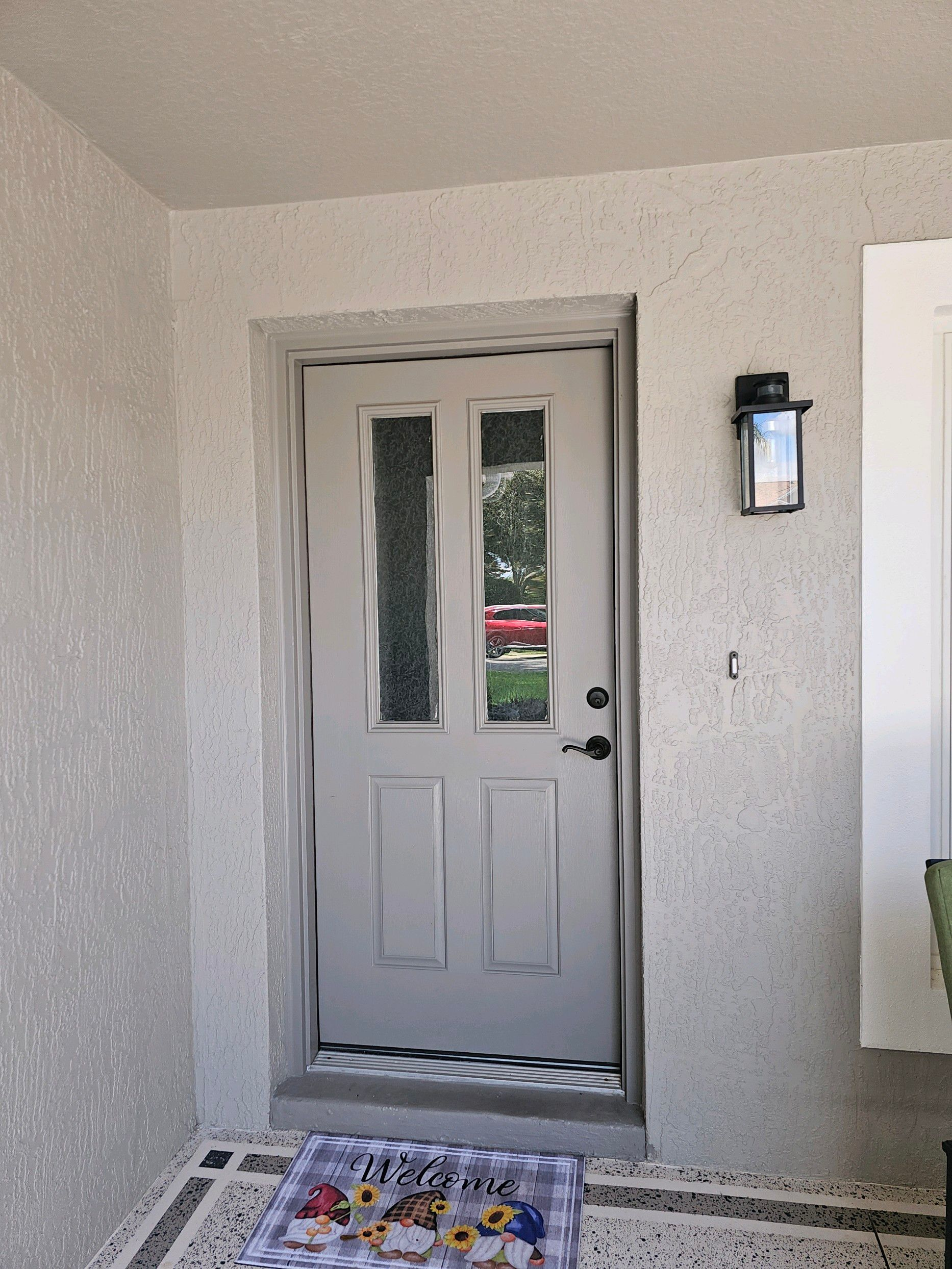 Gray front door with glass panels, outdoor lamp, welcome mat. Light-colored textured walls.