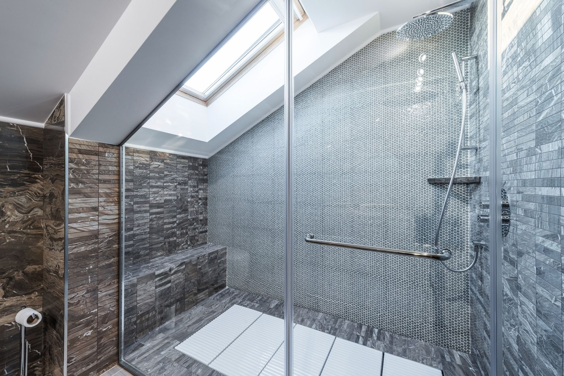 Shower with glass door, gray tiled walls, skylight, white bath mat.