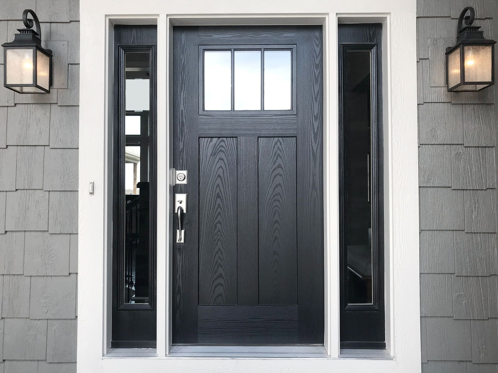 Black front door with sidelights, under a white frame, with gray siding and lanterns.
