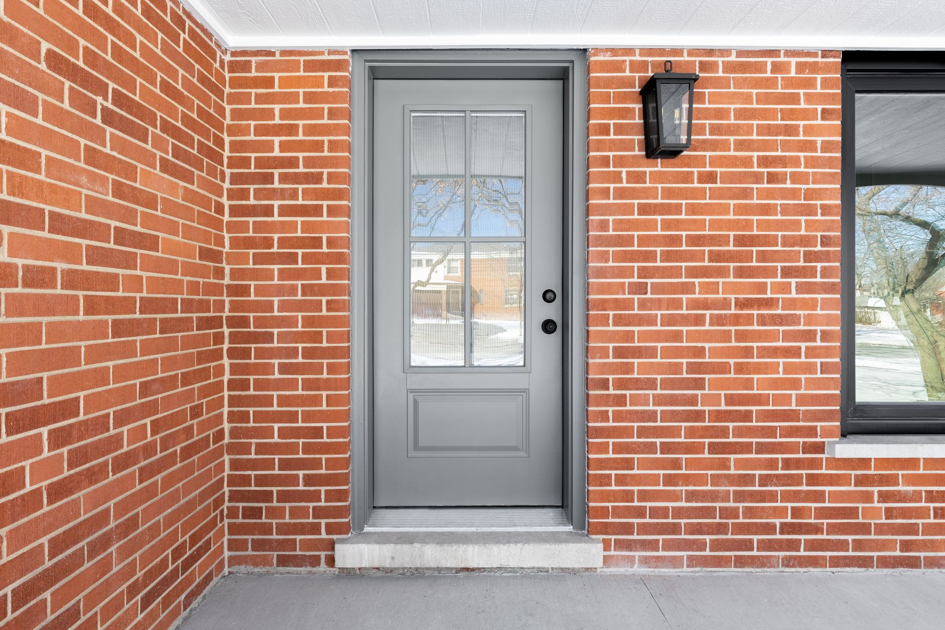 Gray front door with glass panel on red brick wall; black sconce and window.