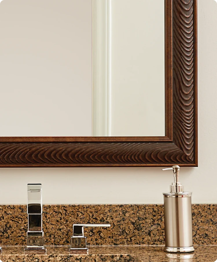 Mirror with textured wood frame above a bathroom sink with chrome fixtures and a lotion dispenser.