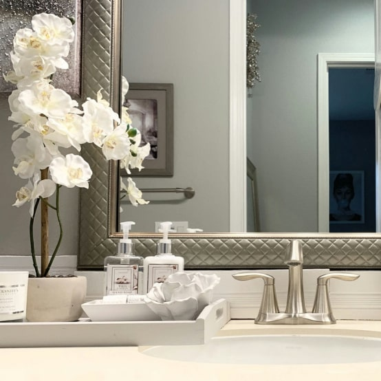 Bathroom countertop with white orchids, soap dispensers, tray, and a silver-framed mirror.