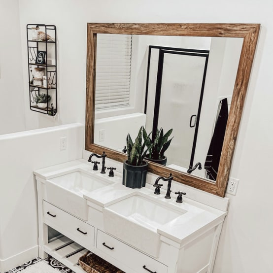 Bathroom with a white double vanity, farmhouse sinks, black fixtures, and a large wooden mirror.