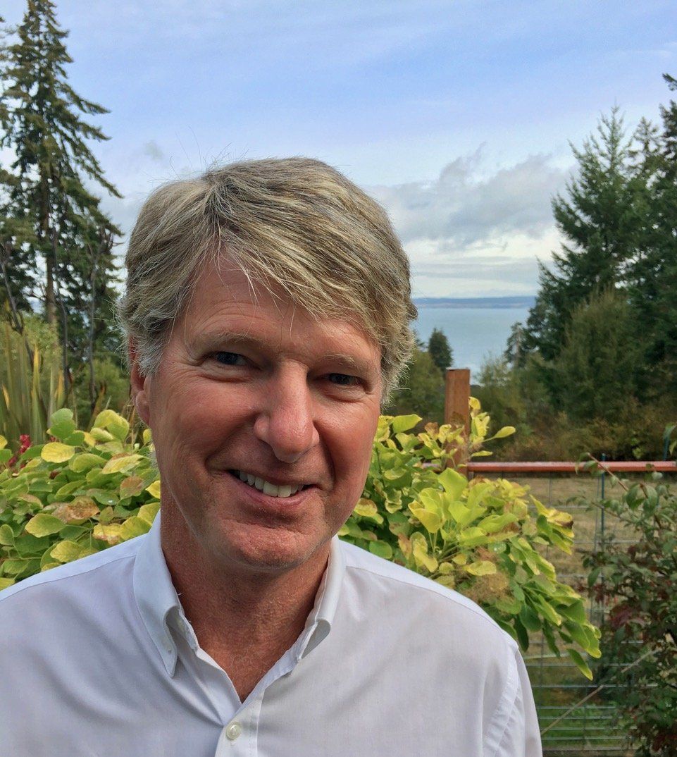 A man in a white shirt is smiling in front of trees and a body of water.