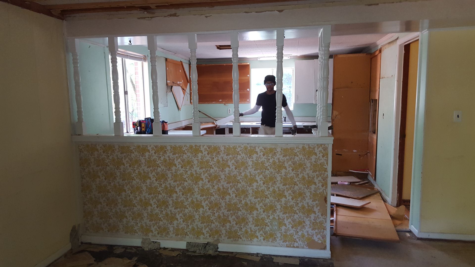 A person stands in a kitchen visible behind a half-wall featuring yellow patterned wallpaper and white spindles.