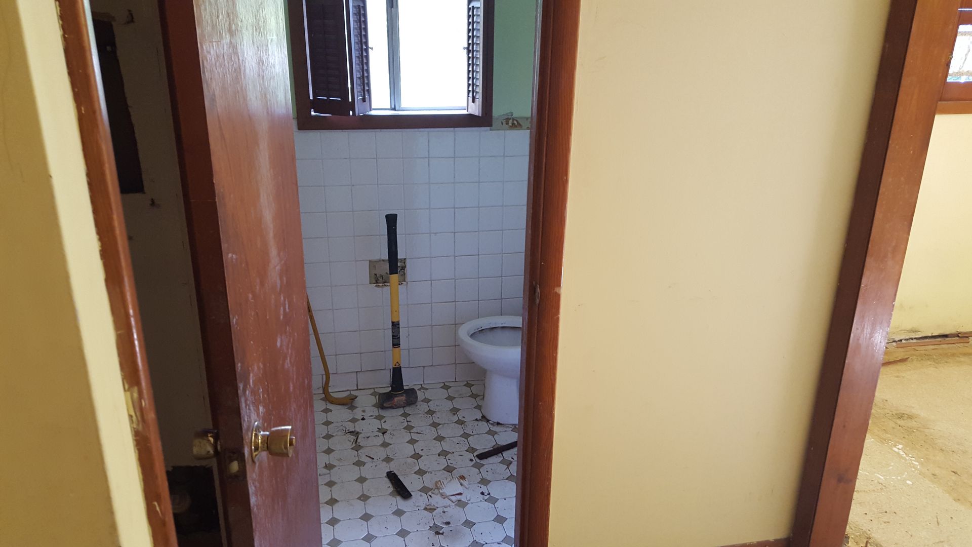 A view through an open doorway into a small, tiled bathroom containing a white toilet and a long-handled floor scraper.