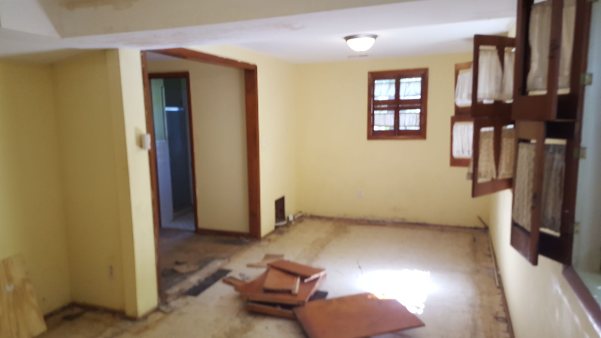 An interior view of a room with yellow walls, a window, a doorway leading to another room, and loose floor tiles.