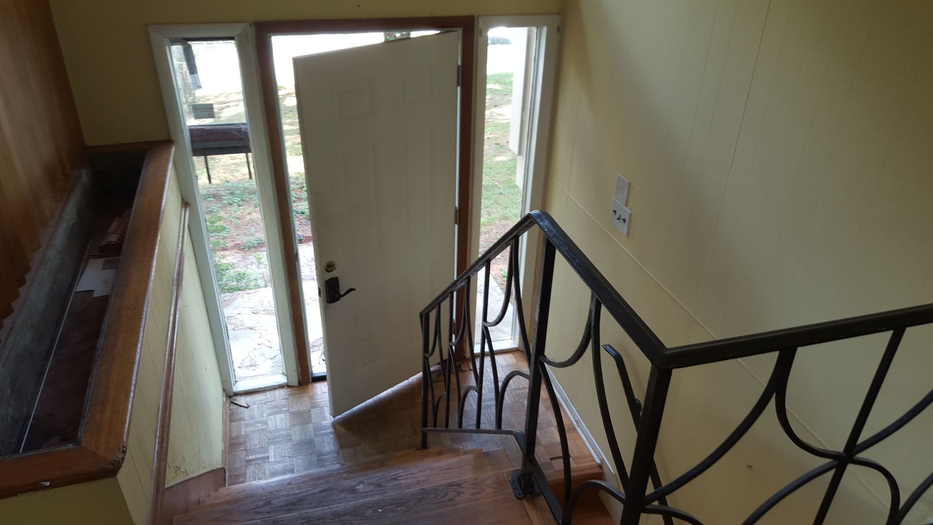 A view looking down a wooden staircase toward an open white front door with glass sidelights and a black metal railing.