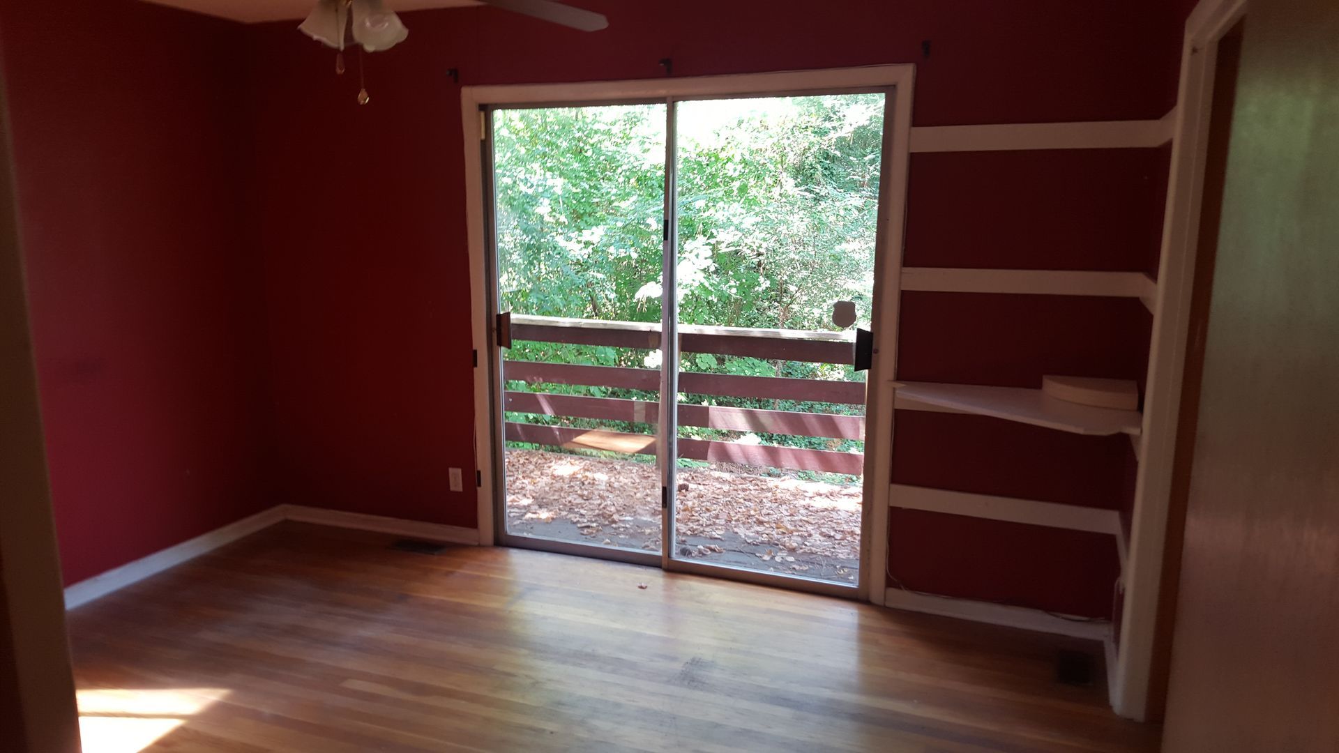 A room with red walls, hardwood floors, a sliding glass door leading to a balcony, and built-in white shelves.