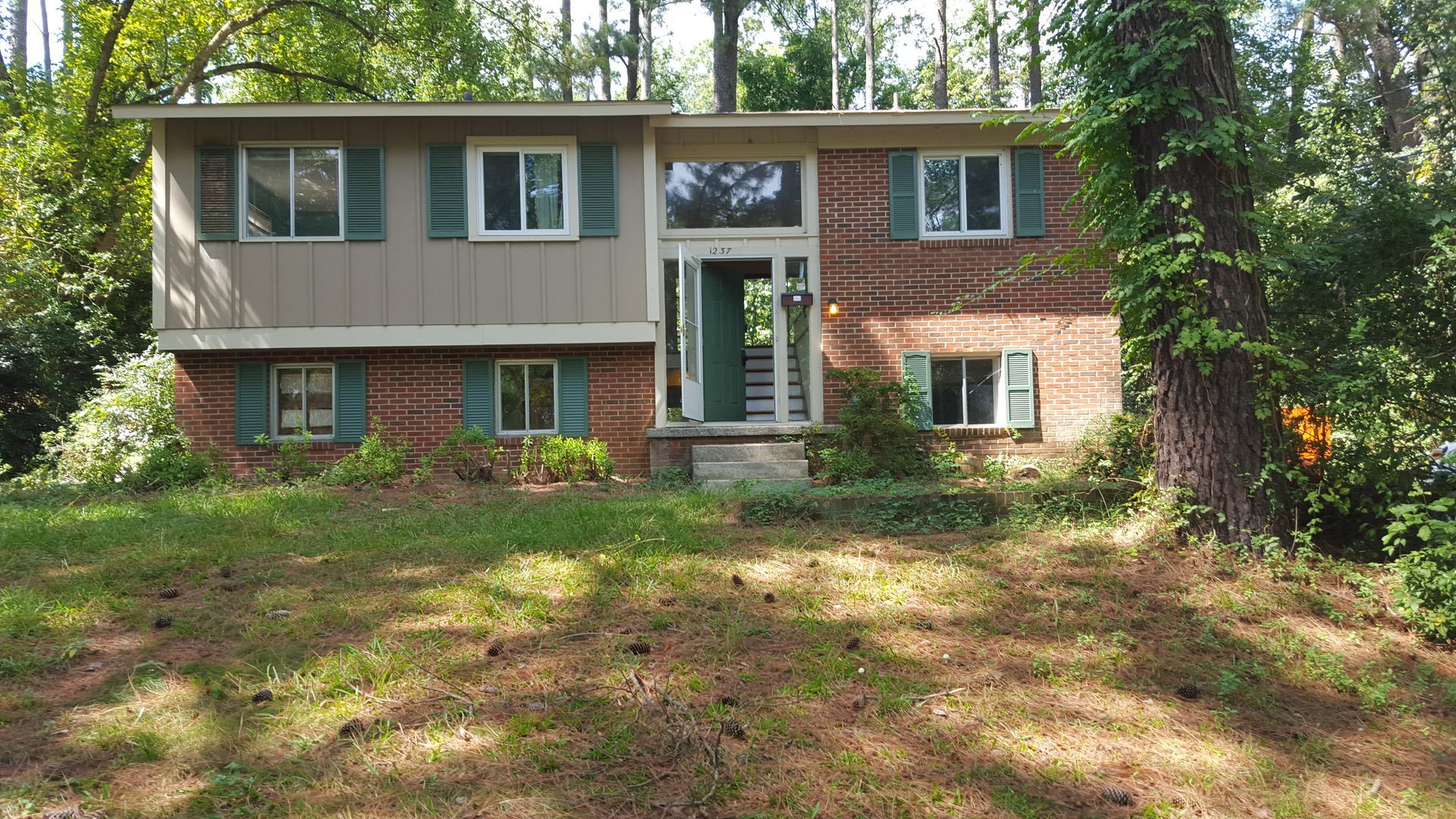 A two-story split-level home with brick and tan siding, green shutters, and a central entryway, surrounded by trees.