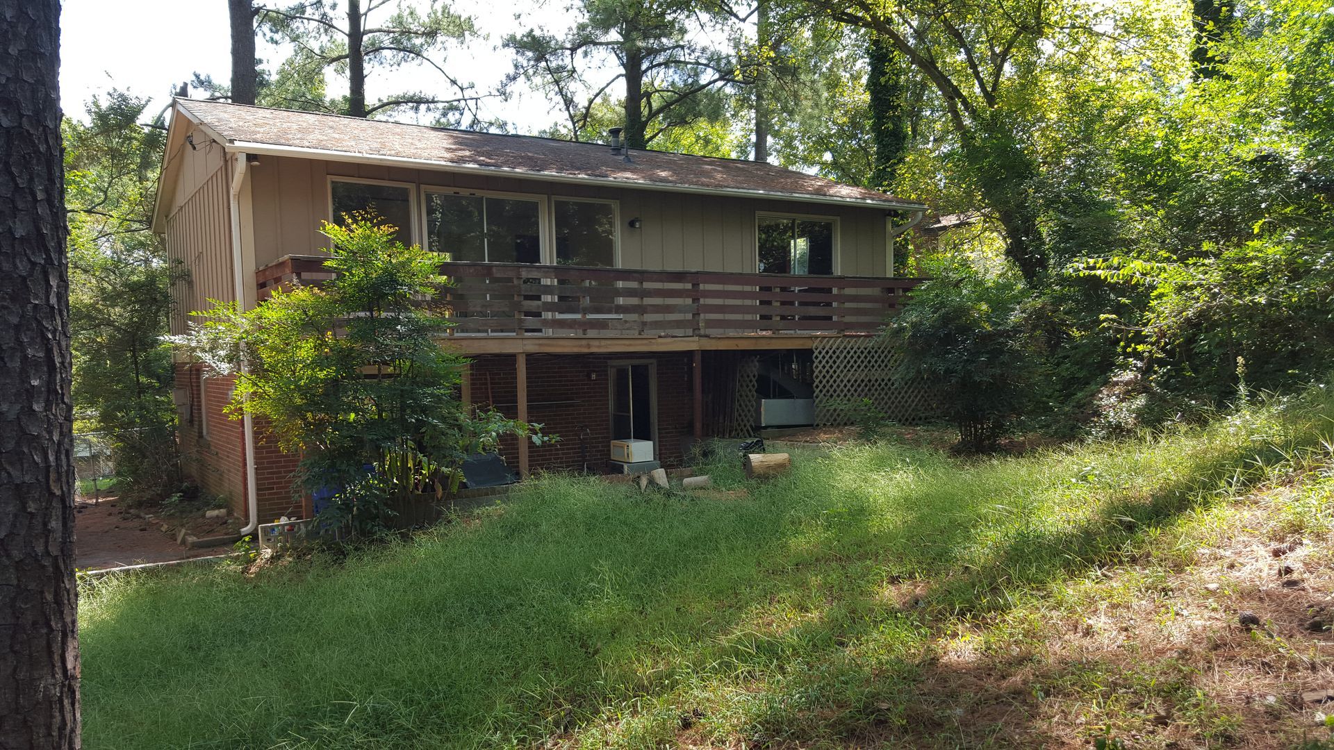 A tan house with a wooden deck nestled in a wooded, grassy hillside.